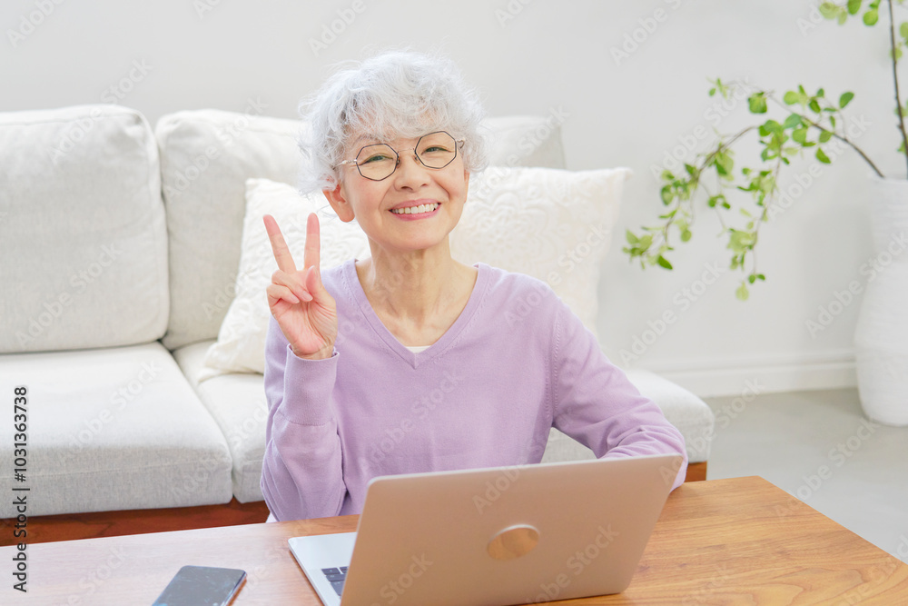 ノートパソコンの前でピースサインするシニア女性 リビングルーム ノートパソコンの前でピースサインするシニア女性 リビングルーム