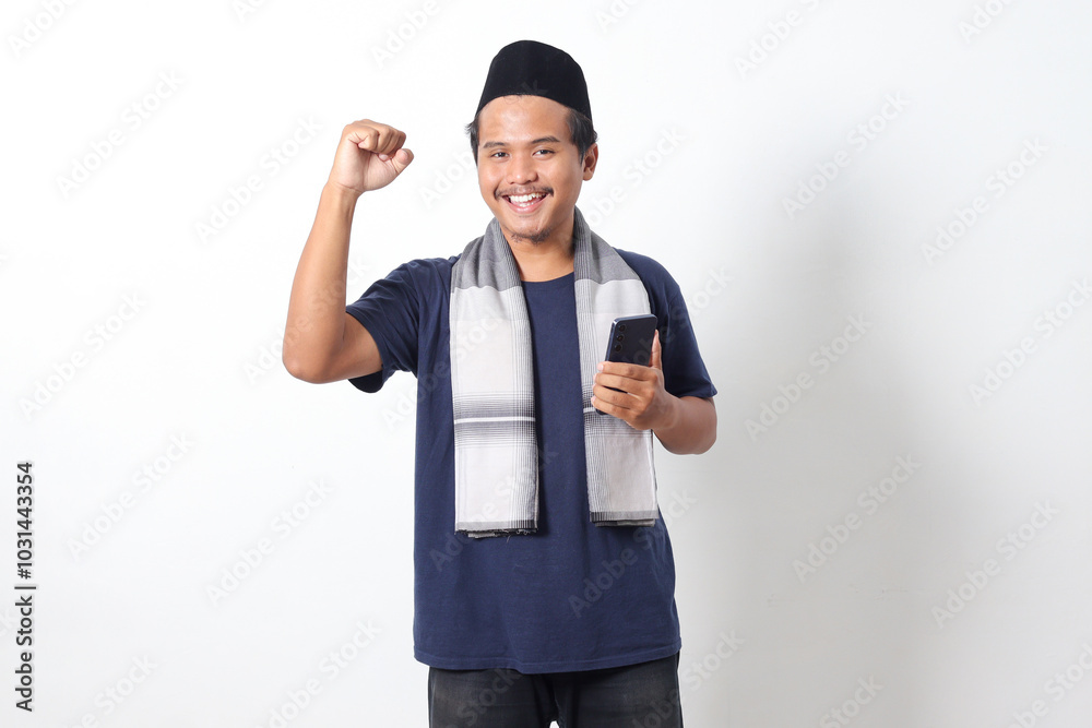 Fototapeta premium Portrait of excited Asian muslim man wearing sarung scarf and skullcap, raising his fist, celebrating winning game or getting good news on his mobile phone. Isolated on white background