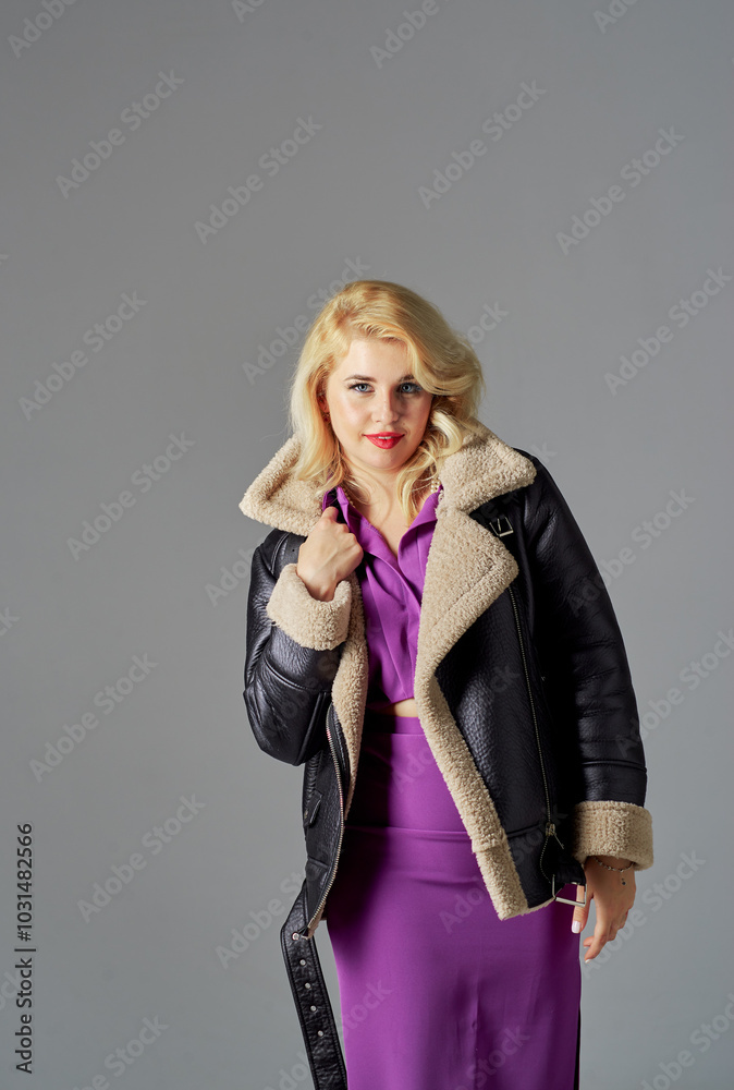 A beautiful girl with blond hair in a purple dress stands on a gray background. She is wearing a black jacket with a fur collar. A beautiful girl with blond hair in a purple dress stands on a gray background. She is wearing a black jacket with a fur collar.