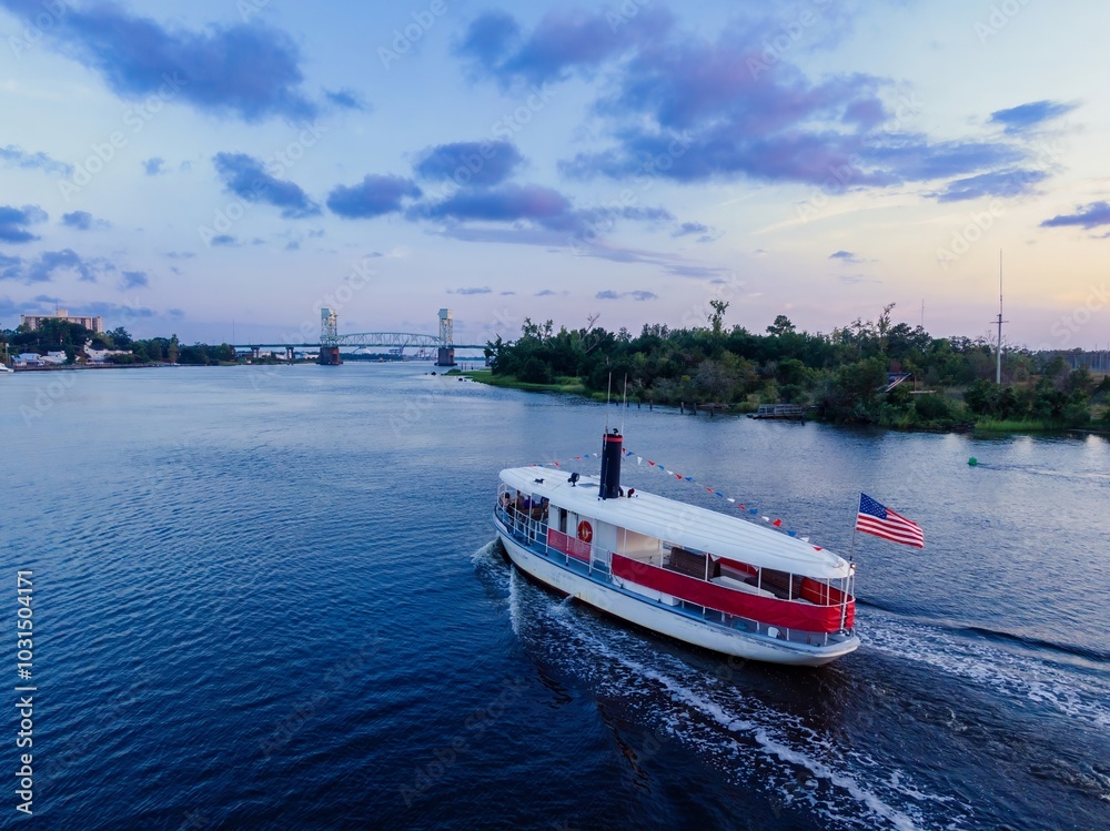 Obraz premium Tourist ferry boat on the Cape Fear River and the Cape Fear Memorial Bridge at sunset in Wilmington, North Carolina, United States.