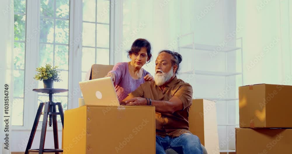 Indian Asian middle-aged married couple sitting on the floor of their new home surrounded by moving boxes, using a laptop to work on interior design plans, happily settling into their new apartment Indian Asian middle-aged married couple sitting on the floor of their new home surrounded by moving boxes, using a laptop to work on interior design plans, happily settling into their new apartment