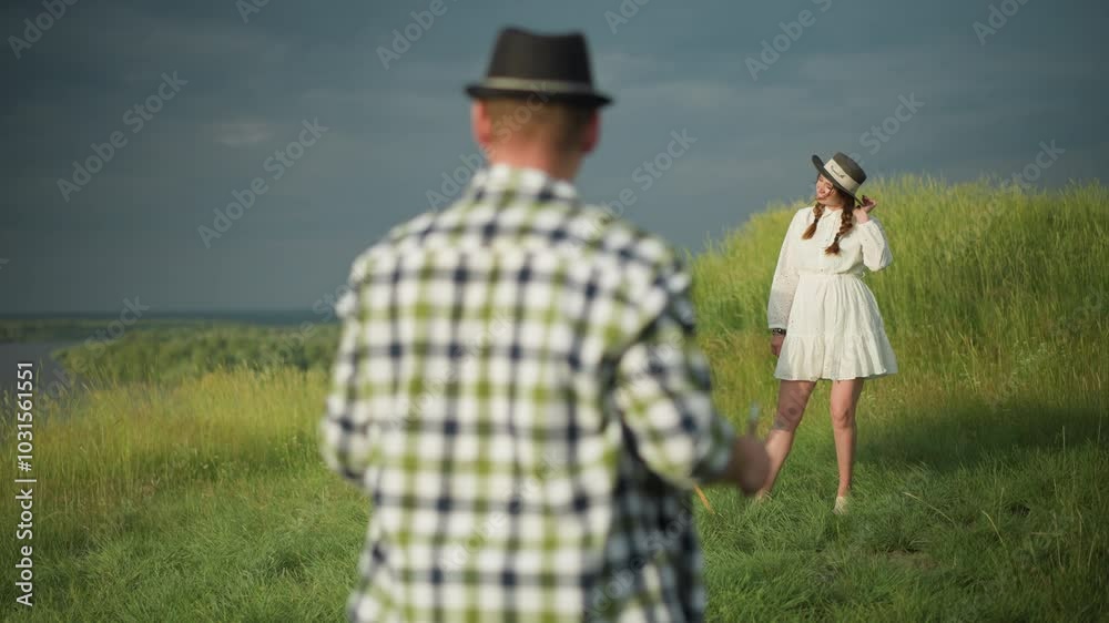 A woman wearing a white dress and hat stands in front of a canvas in a grassy field, striking a pose as a man in a checkered shirt approaches her. The scene is set in a serene outdoor environment A woman wearing a white dress and hat stands in front of a canvas in a grassy field, striking a pose as a man in a checkered shirt approaches her. The scene is set in a serene outdoor environment