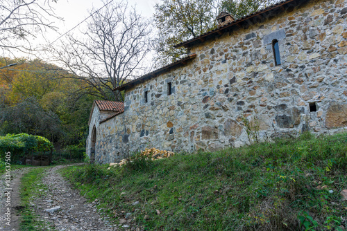 A gravel road leads to the outer wall and gate of the monastery. A hill covered with autumn trees is in the background.