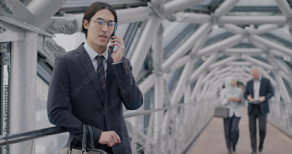 Asian businessman speaking on mobile phone standing in unique glass hall while people in formalwear walking in hallway. Communication and business concept. Asian businessman speaking on mobile phone standing in unique glass hall while people in formalwear walking in hallway. Communication and business concept.