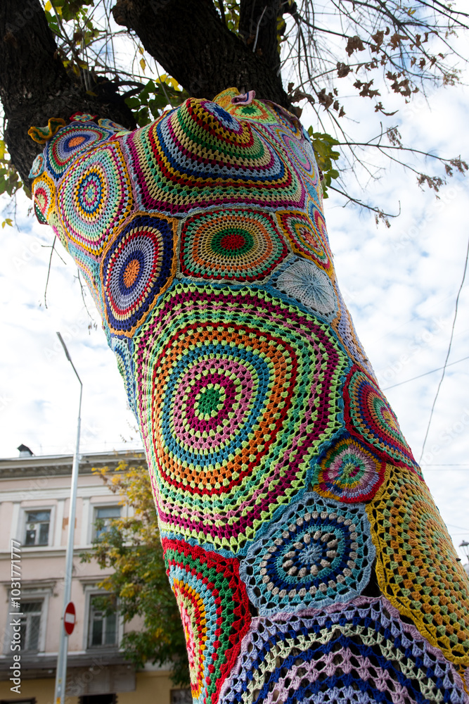 Naklejka premium A tree covered in colorful crocheted patterns as part of urban yarn bombing, with vibrant geometric shapes and textures. The creative installation stands out against the natural and urban backdrop
