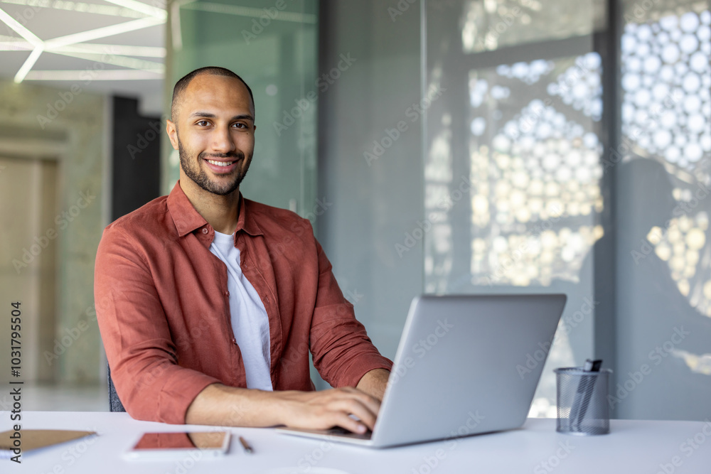 Smiling man working on laptop at desk in modern office, showcasing productivity. Table holds smartphone and pen holder, reflecting efficiency and work environment. Bright, professional setting. Smiling man working on laptop at desk in modern office, showcasing productivity. Table holds smartphone and pen holder, reflecting efficiency and work environment. Bright, professional setting.