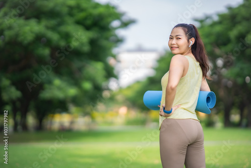 Young Asian Beautiful woman in yellow armless t-shirt and brown legging holding roll yoga mat wearing ear bud and smartwatch looking at camera with smile of peacful and happiness.