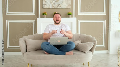 successful smiling bearded man in white t shirt and jeans sitting on sofa with laptop at home in room