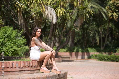 Free Download A young, dark-haired, beautiful woman, heavily tattooed, in a white dress, sitting on a stone bench with a serious and defiant look. Tattoos concept, nonconformism, rebellious, trend, fashion. - GFXLoad