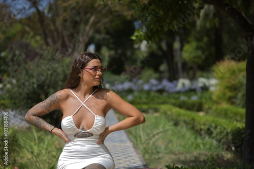 A young, dark-haired, beautiful woman, heavily tattooed, in a white dress and sunglasses with purple crystals, sitting, with her hands on her waist, looking into infinity relaxed and calm.