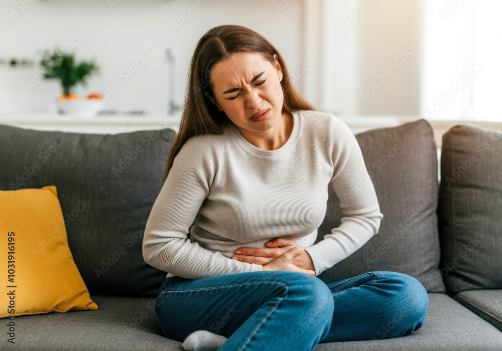 Young woman experiencing abdominal pain while sitting on a sofa in a bright, cozy living room during the afternoon Young woman experiencing abdominal pain while sitting on a sofa in a bright, cozy living room during the afternoon