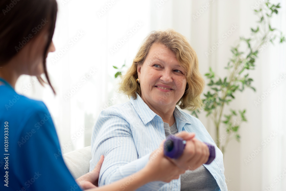 Obraz premium Asian caregiver woman or nurse training senior woman lifting dumbbell for arm exercise while therapy and rehabilitation in living room at home, caretaker or physiotherapist helping elderly workout.