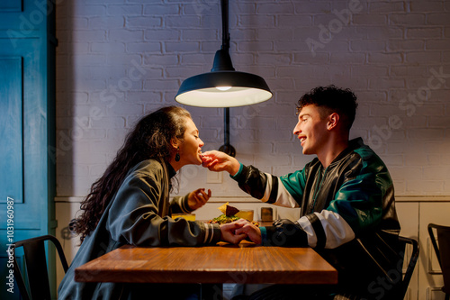 Young white man having dinner with his Latina girlfriend. Multiracial couple having dinner together. Boyfriend shoves a portion of Mexican nachos into his girlfriend's mouth while eating at a restaura photo