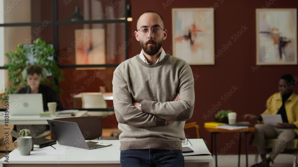 Portrait of Caucasian young man with crossed arms posing for camera in modern office near work desk, two colleagues in background Portrait of Caucasian young man with crossed arms posing for camera in modern office near work desk, two colleagues in background