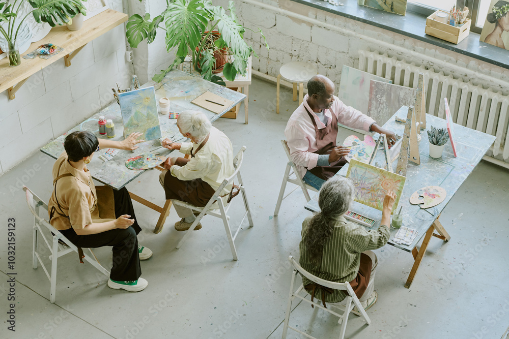 High angle shot of retro drawing studio with biracial people of different age sitting at work desks and painting on canvas High angle shot of retro drawing studio with biracial people of different age sitting at work desks and painting on canvas