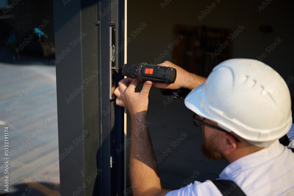 Construction worker installing door in a new house construction site. Man in working attire install front door. Builder in uniform with screwdriver adjusting front glass door in new beautiful house. Construction worker installing door in a new house construction site. Man in working attire install front door. Builder in uniform with screwdriver adjusting front glass door in new beautiful house.