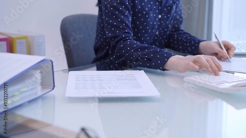 A female accountant with blue dotted blousy is using a laptop computer and calculator to calculate taxes at a glass desk in the office. Right side view photo