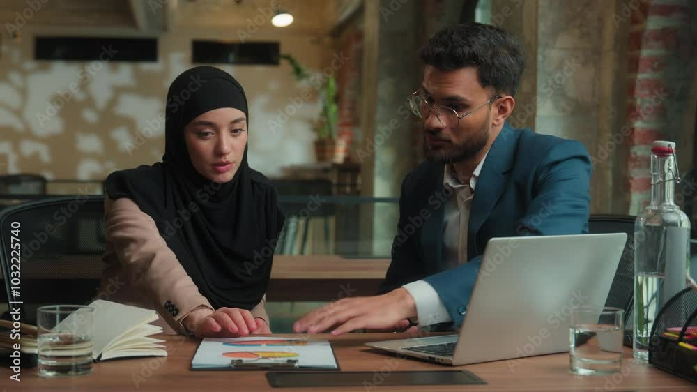 Two multiethnic business partners colleagues male female workers man muslim woman in hijab talk discuss marketing graphs analyzing documents on table office Arabian Indian businessman businesswoman