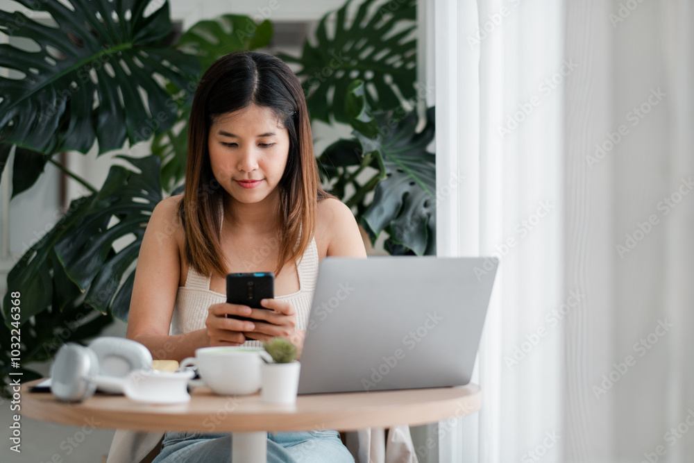 A young woman sits at a table using her smartphone and laptop, surrounded by green plants in a modern home office setting. A young woman sits at a table using her smartphone and laptop, surrounded by green plants in a modern home office setting.