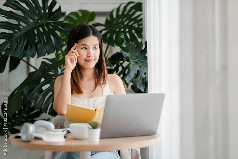 A young woman sits at a table with a laptop and notebook, working from home in a bright, modern room with large green plants. A young woman sits at a table with a laptop and notebook, working from home in a bright, modern room with large green plants.