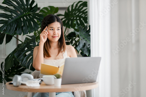 A young woman sits at a table with a laptop and notebook, working from home in a bright, modern room with large green plants.