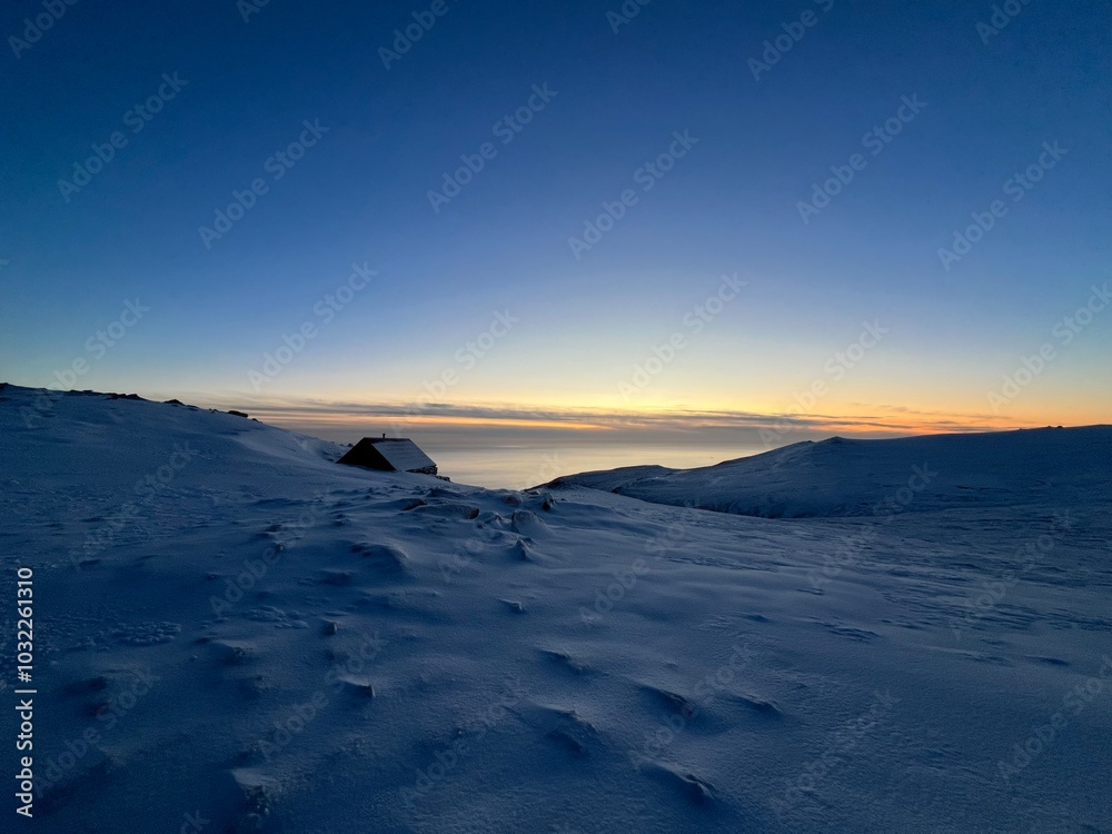 Obraz premium Seaside winter view. The photo captures a peaceful, snowy landscape at dusk. A small, dark cabin sits on the left side, partially buried in snow, with its roof covered in frost.
