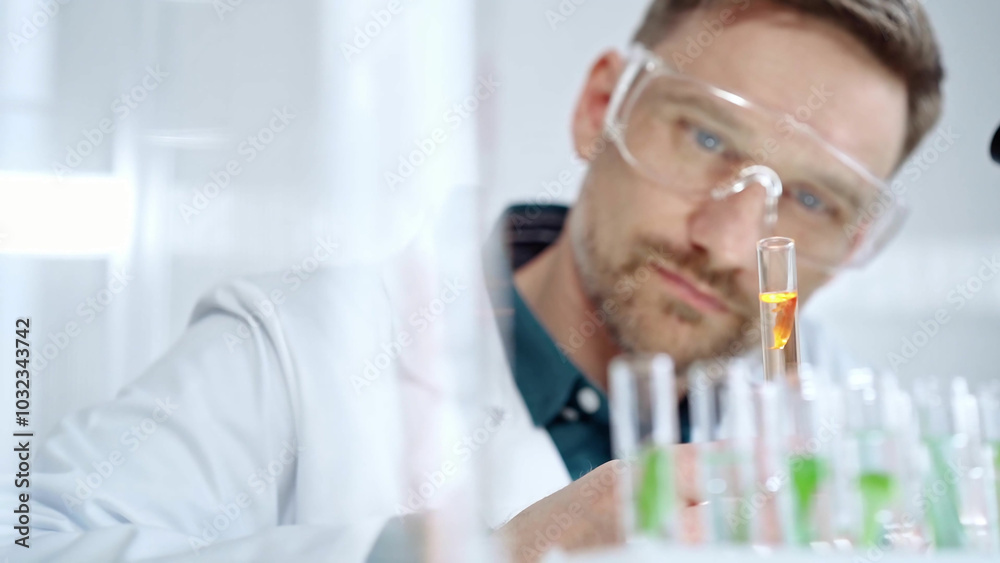 Man researcher with safety glasses is examining lab tube with a orange fluid in a modern laboratory, close-up. Science, microbiology and medicine Man researcher with safety glasses is examining lab tube with a orange fluid in a modern laboratory, close-up. Science, microbiology and medicine