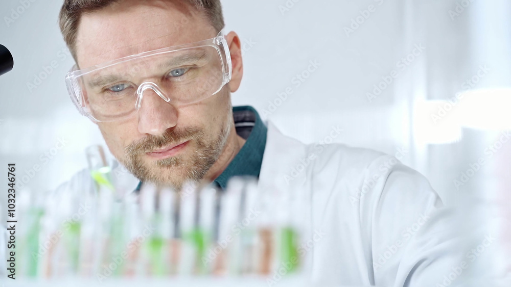 Man researcher with safety glasses is examining lab tube with a green fluid in a modern laboratory, close-up. Science, microbiology and medicine Man researcher with safety glasses is examining lab tube with a green fluid in a modern laboratory, close-up. Science, microbiology and medicine