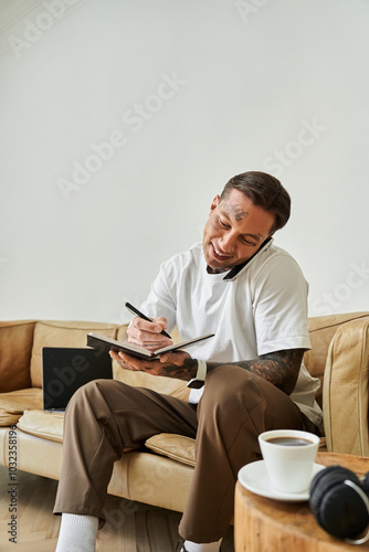 A young man sits comfortably at home, engaging in phone conversations and taking notes.