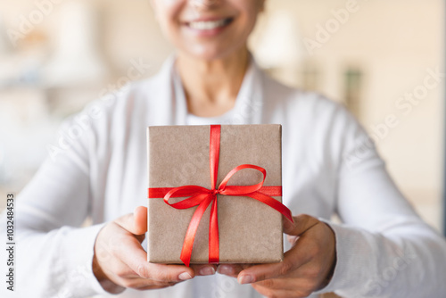 Mature smiling beautiful woman holding focused present gift box with red ribbon, giving receiving presents on holiday event, celebration Christmas birthday at home