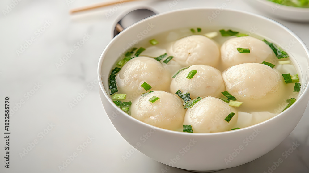 A bowl of soup with dumplings, garnished with chopped green onions, placed on a white table with chopsticks and a spoon nearby. A bowl of soup with dumplings, garnished with chopped green onions, placed on a white table with chopsticks and a spoon nearby.