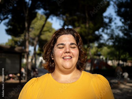 A young woman with intellectual disability smiles while receiving the sun at the face with closed eyes, standing outdoors in a park. The sun shines brightly, and trees surround her, creating a joyful  photo