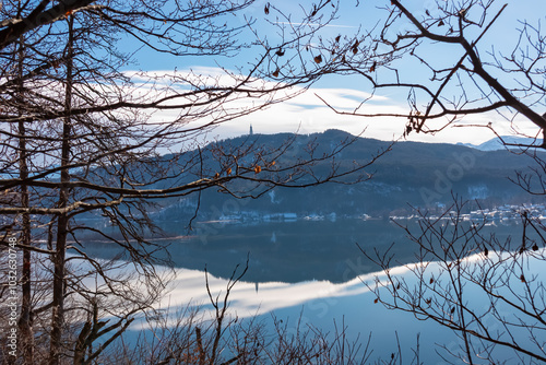 Scenic view of alpine Lake Wörth surrounded by snow-capped mountains Karawanks in Carinthia, Austria, Europe. Viewing tower Pyramidenkogel reflected in calm water surface. Hohe Gloriette, Pörtschach