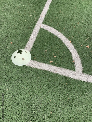 A close-up shot of a corner area on a green sports field marked with white lines, most likely a soccer field. A light green plastic training cone is placed near the corner line.