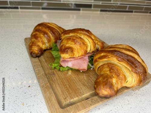 A delicious croissant sandwich filled with ham and lettuce, presented on a wooden cutting board in a modern kitchen setting. A medium-angle shot with a neutral background.