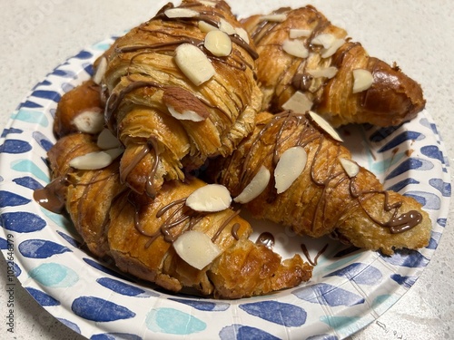 Croissants topped with sliced almonds and drizzled with chocolate, displayed on a patterned paper plate. A perfect treat for dessert or breakfast.