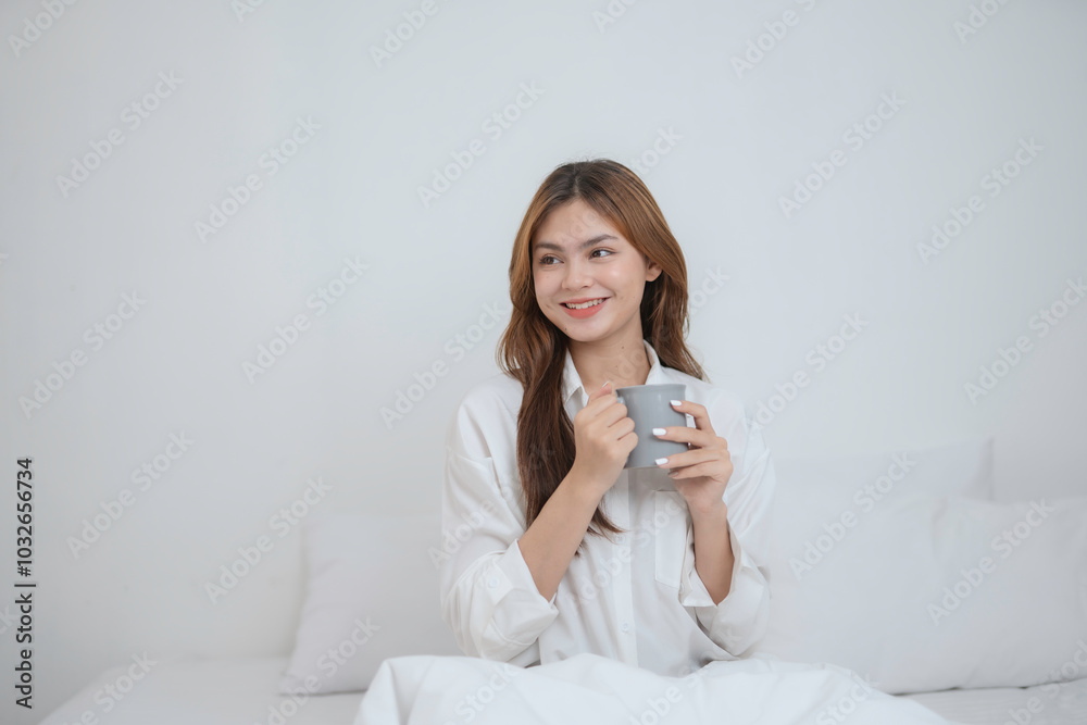 Young woman sits up in bed, smiling with a warm mug in hand, surrounded by cozy bedroom light. Her brunette hair falls gently, exuding comfort and relaxation