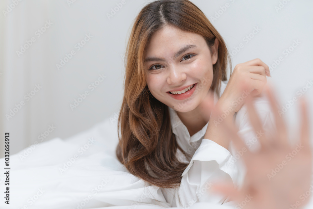 Beautiful young asian woman with long brown hair is lying in bed with white sheets and a white shirt, smiling and raising her hand Beautiful young asian woman with long brown hair is lying in bed with white sheets and a white shirt, smiling and raising her hand