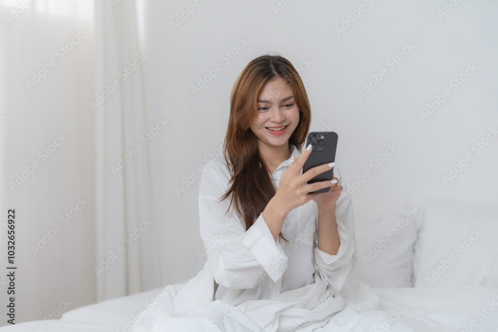 Young woman is sitting in bed with a smile on her face, holding a smartphone in her hands and browsing social media Young woman is sitting in bed with a smile on her face, holding a smartphone in her hands and browsing social media