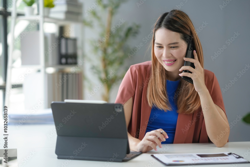 Asian businesswoman is working in her bright office, looking at a tablet and smiling while talking on the phone Asian businesswoman is working in her bright office, looking at a tablet and smiling while talking on the phone