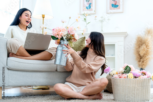 Young Asian woman show her success of flower rearrangement to her sister who sit on sofa with laptop and they look happy together. photo