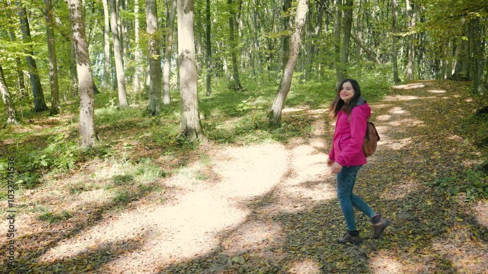 Attractive young woman in a pink jacket, jeans, and brown backpack walking through an autumn forest, turns, smiles at camera and gestures follow me or let's go with her hand, slow motion Attractive young woman in a pink jacket, jeans, and brown backpack walking through an autumn forest, turns, smiles at camera and gestures follow me or let's go with her hand, slow motion