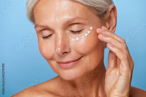 Smiling mature woman with closed eyes applying moisturiser under her eyes, showing a relaxed skincare routine against a blue background... photo