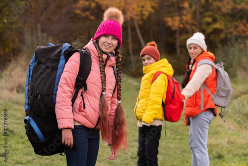 family walking in autumn forest with backpacks, boy in yellow jacket, girl in pink jacket, mother in orange sleeveless jacket, family in hat with pompoms, gen z photo