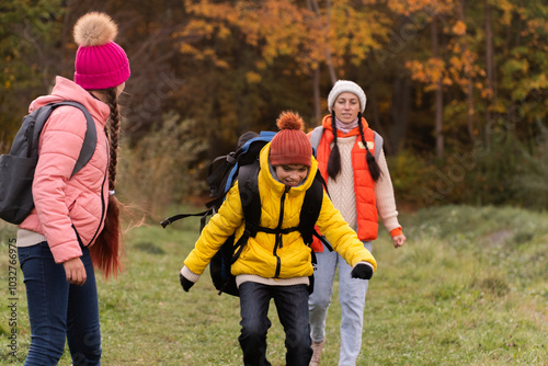 family walking in autumn forest with backpacks, boy in yellow jacket, girl in pink jacket, mother in orange sleeveless jacket, family in hat with pompoms, gen z