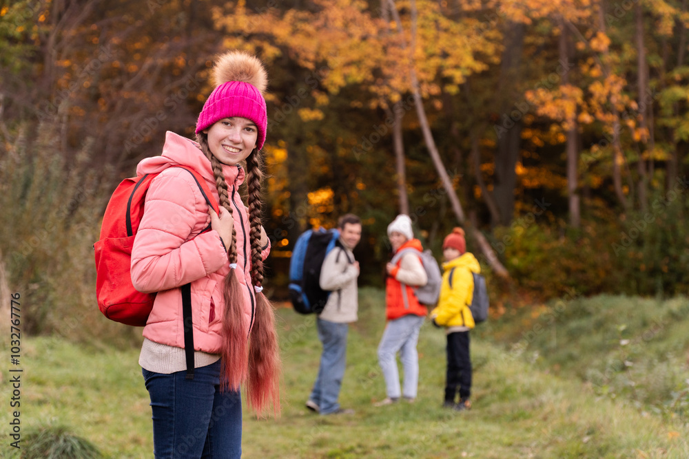 family walking in autumn forest with backpacks, boy in yellow jacket, girl in pink jacket, mother in orange sleeveless jacket, family in hat with pompoms, gen z, father with a big backpack