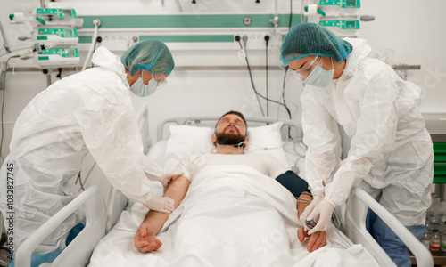 Fotografija A male patient lies in a hospital bed with an oxygen mask, while two healthcare workers in full protective suits and masks attend to him in a modern medical facility