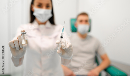 Masked healthcare professional holds up syringe and vial of vaccine, while a masked male patient waits in the background. The image emphasizes vaccination and healthcare safety in a clinical setting