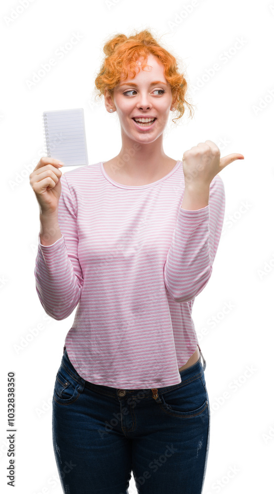 Young redhead woman holding blank notebook pointing and showing with thumb up to the side with happy face smiling Young redhead woman holding blank notebook pointing and showing with thumb up to the side with happy face smiling