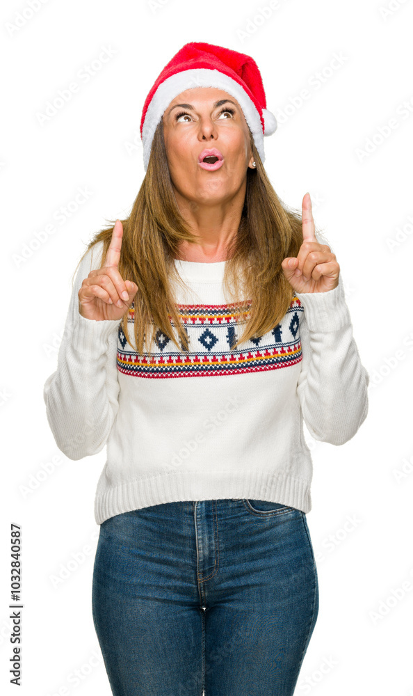 Middle age adult woman wearing winter sweater and chrismat hat over isolated background amazed and surprised looking up and pointing with fingers and raised arms. Middle age adult woman wearing winter sweater and chrismat hat over isolated background amazed and surprised looking up and pointing with fingers and raised arms.